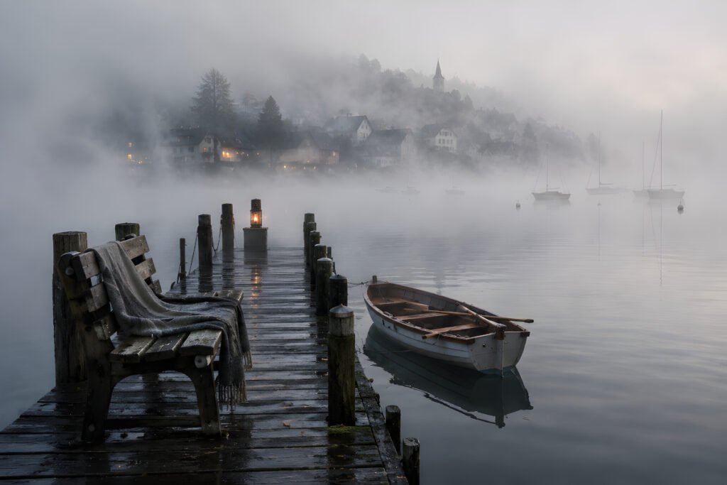 The Fog that Carried Voices – weathered wooden dock and rowing boat surrounded by thick morning mist on a quiet lake.