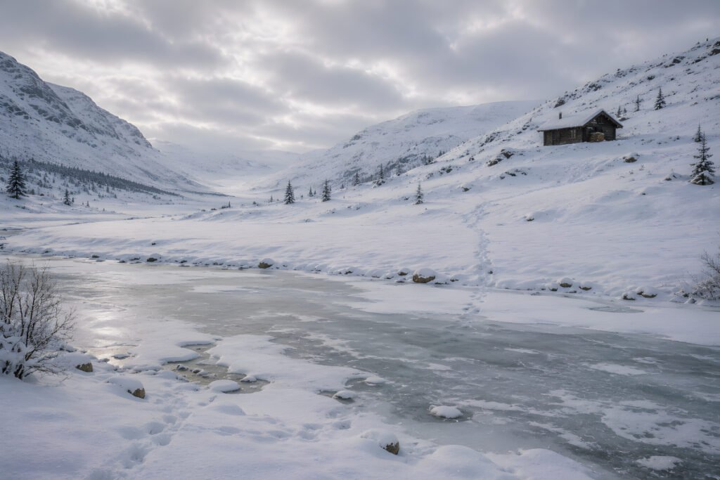 The Hut Across the Snow Valley – a frozen river crosses the snowy valley below a solitary wooden hut standing on the rising slope beyond.